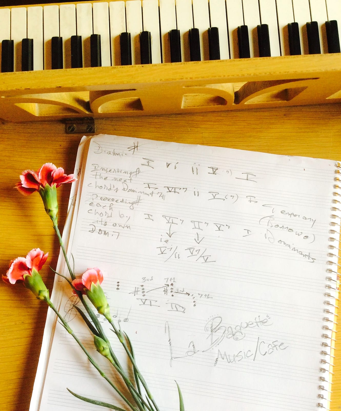 Handwritten music theory notes in a spiral notebook on a wooden piano, with pink carnations resting beside it